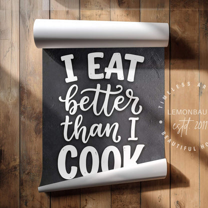 A rolled-up black sign with white lettering that reads 'I EAT better than I COOK' displayed on a rustic wooden plank background.